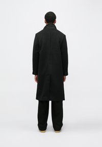 Sort uldfrakke med lige snit, lang længde og høj krave; vist bagfra, kombineret med sorte bukser og lysebrune sko.