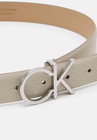 Cintura in pelle grigia chiara con una fibbia circolare in ottone argento con logo CK e multiple aperture di regolazione per flessibilità nelle misure.