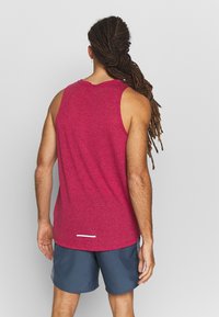 Rotes ärmelloses Top mit einem strukturierten Muster, abgerundetem Saum und einem reflektierenden Detail am unteren Rand. Getragen mit marineblauen Shorts.