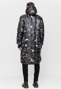 Pikowana czarna kurtka z błyszczącym wykończeniem, z białym wzorem rozprysków, długiego kroju i odpinanym kapturem. Noszone czarne spodnie i buty.