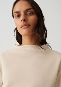 Top beige a collo alto con texture liscia simile alla satin, caratterizzato da un design minimalista e senza motivi o accessori visibili.