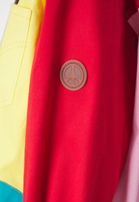 Colorful jacket featuring red and yellow fabric, with a brown circular peace symbol patch. Smooth texture and functional design elements.