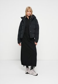 Svart puffig jacka med huva, kort design, i kombination med en lång svart kjol och ljusa sneakers, på en neutral bakgrund.