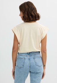 Top sin mangas de color crema con hombros enrollados, combinado con jeans de mezclilla azul de tiro alto. Tejido suave con una textura lisa.