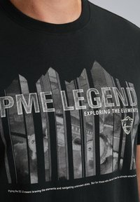 Čierne bavlnené tričko s grafickým dizajnom a bielym textom "PME LEGEND" a "EXPLORING THE ELEMENTS." Textúrovaný povrch so pruhmi.