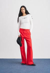 Baltas ilgomis rankovėmis viršus su raudonomis detalėmis, derinamas su raudonomis sportinėmis kelnėmis su elastinga juosmens juosta. Juoda kepurė rankoje. Lygus, tamprus audinys.