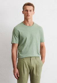 Green short-sleeve t-shirt made of lightweight fabric, featuring a round neckline. Worn with light green linen pants, showing relaxed fit.