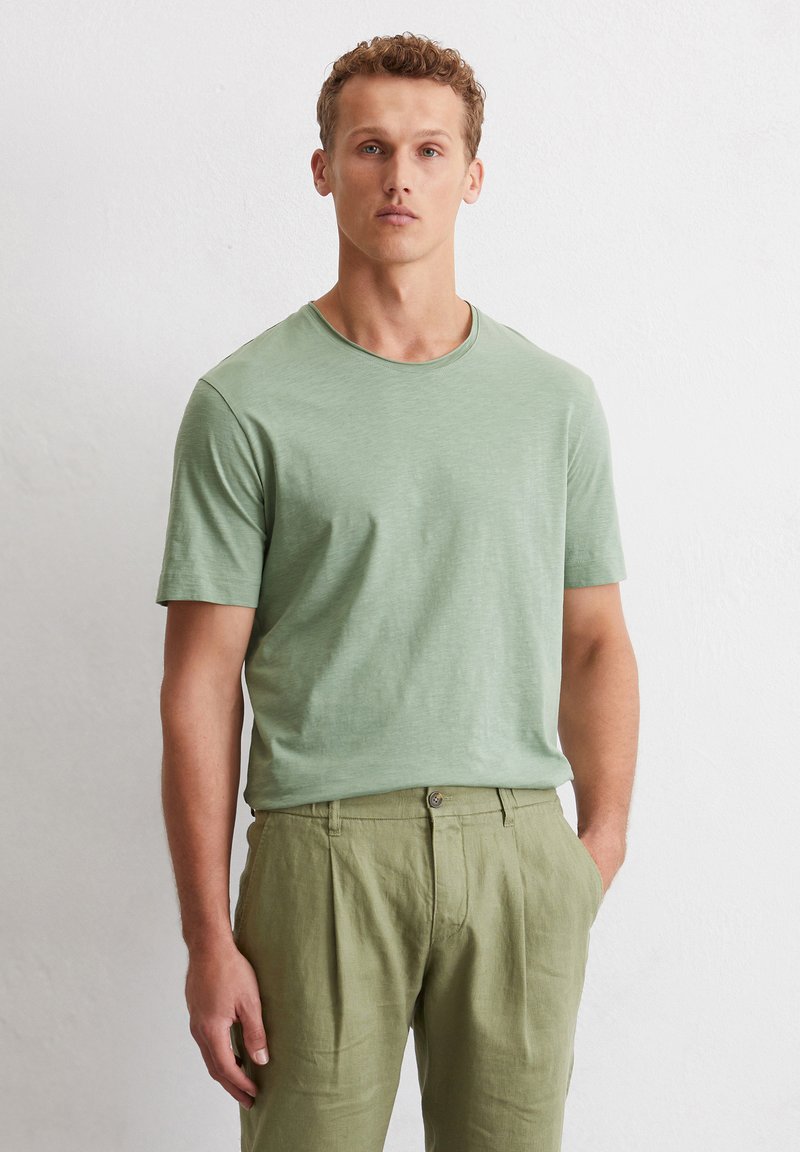 Green short-sleeve t-shirt made of lightweight fabric, featuring a round neckline. Worn with light green linen pants, showing relaxed fit.