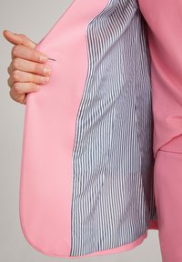 Käsi, der hält einen offenen pinken Blazer, der eine gestreifte blaue und weiße Futter zeigt, vor einem schlichten Hintergrund.