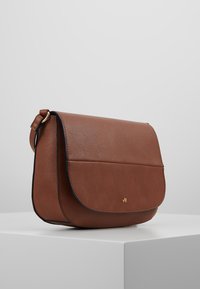 Hnědá kožená kabelka přes rameno s klopou, zaobleným tvarem, zlatými kovovými prvky a kontrastním černým prošíváním. Hladký povrch.