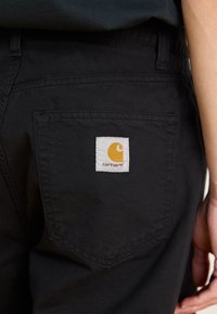 Black pants featuring a back pocket with a visible white label displaying an orange logo. Material appears sturdy with a smooth texture.