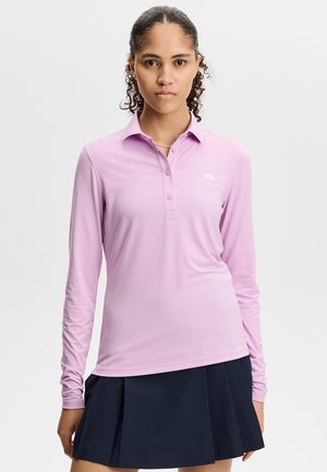 Vrouw met krullend haar vastgebonden draagt een lichtpaarse lange mouwen poloshirt en een zwarte geplooide rok tegen een effen witte achtergrond.