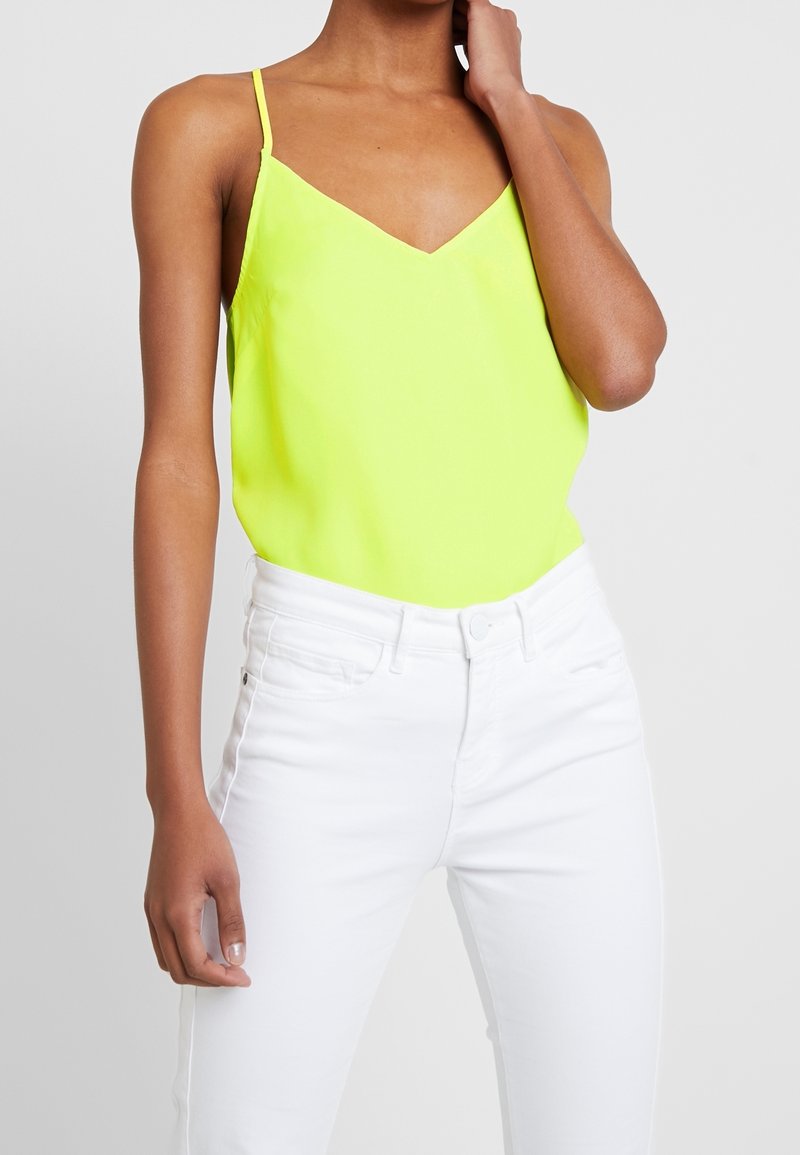 Top senza maniche giallo neon con scollo a V e spalline sottili, abbinato a jeans bianchi aderenti. Texture liscia, design semplice, senza motivi visibili.