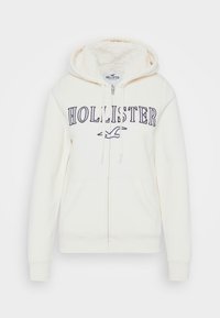 Vit zip-up hoodie med en mjuk, fodrad insida, med "Hollister" i marinblått på framsidan, två sidofickor och ribbade ärmslut.