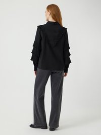 Svart skjorte med rysjet erme og V-formet krage bak, kombinert med grå vidbukser og svarte sko, på lys bakgrunn.