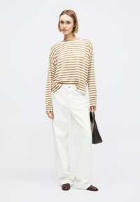 Femme aux cheveux blonds courts portant un chemisier à manches longues rayé marron et blanc, un pantalon large blanc, des chaussures marron, tenant un sac à main noir.