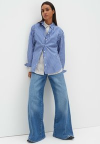 Blau-weiß gestreiftes Hemd mit Knöpfen, darüber getragen ein weißes und schwarz gestreiftes Shirt, kombiniert mit weit geschnittenen Jeans.