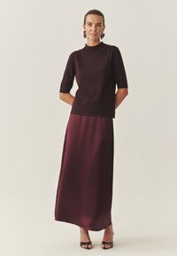 Robe bordeaux mi-longue avec un haut ajusté et des manches courtes, présentant une texture lisse et brillante. Assortie à des talons ouverts foncés.