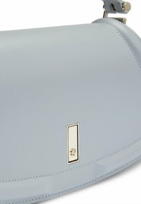 Lysblå lædertaske med en glat overflade, som har en guldfarvet rektangulær hardwaredetalje og en buet form. Minimalistisk design.