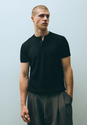 Jeune homme aux cheveux courts portant une chemise noire à manches courtes boutonnée et un pantalon gris, debout avec une main dans la poche.