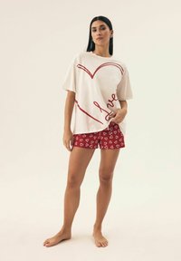Biały, oversized T-shirt z grafiką czerwonego serca i napisem "love", w połączeniu z czerwonymi szortami z białymi wzorami serc. Gładki materiał, swobodny styl.