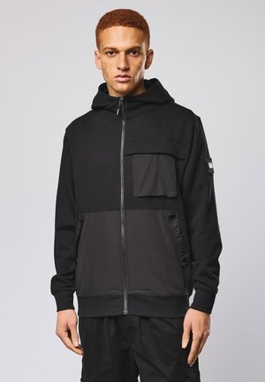 Jonge man met kort rood haar, gekleed in een zwarte hoodie met ritssluiting en grote zakken aan de voorkant en op de borst, tegen een effen lichte achtergrond.