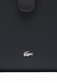 Czarna torebka teksturowa z gładkim wykończeniem, z logo w kolorze srebrnym w kształcie krokodyla i zapięciem na klapkę. Materiał wydaje się trwały i elegancki.