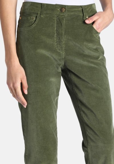 Personne portant un pantalon en velours côtelé vert olive avec les mains reposant près des poches, montrée de la taille à mi-cuisse sur fond blanc.