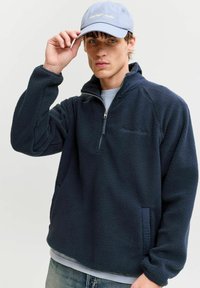 Tânăr ajustând o șapcă albastru deschis, purtând o jachetă fleece bleumarin cu fermoar până la jumătate și o cămașă deschisă la culoare dedesubt, pe fundal neutru.