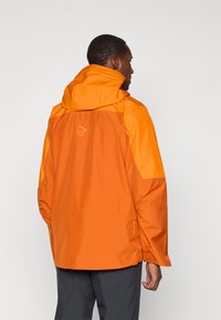 Mies, joka käyttää oranssia huppuvedettävää vedenpitävää takkia, jossa on logo takana, seisoo selkä kameraan päin yksinkertaista vaaleaa taustaa vasten.