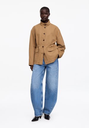 Modelka w beżowej zapinanej na guziki kurtce z dużymi przednimi kieszeniami, szerokich niebieskich jeansach i czarnych butach na obcasie z noskiem w szpic, stojąca na białym tle.