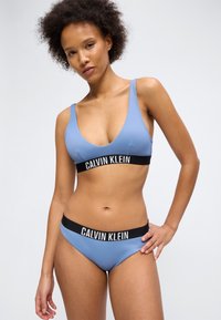 Kobieta o kręconych włosach ubrana w jasnoniebieski biustonosz Calvin Klein i dopasowane majtki z czarnymi elastycznymi taśmami i białym logo.
