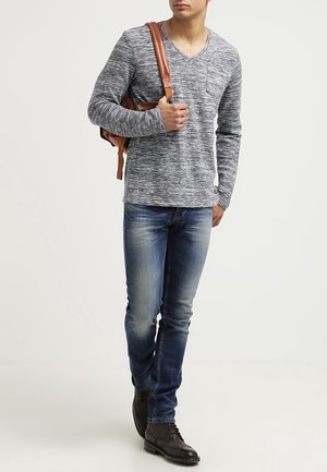 Man draagt een grijs shirt met lange mouwen, blauwe spijkerbroek, bruine nette schoenen, draagt een bruine leren rugzak over één schouder, loopt vooruit.