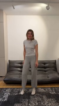 Młoda kobieta w szarej koszulce i dzianinowych spodniach stoi boso na wzorzystym dywanie przed czarną skórzaną sofą w minimalistycznym salonie.