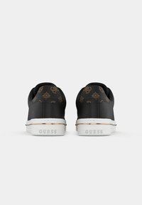 Paire de baskets Guess noires avec des accents marron à motif au talon et des semelles blanches estampillées "GUESS" à l'arrière, vues de dos.