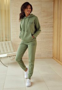 Olijfgroene hoodie en trainingsbroek set, met een voorkant zak, trekkoordcapuchon en bedrukte tekst. Gekoesterd met witte sneakers op een betegelde vloer.