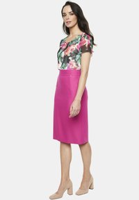 Kwiatowa bluzka z krótkim rękawem w kolorze zielonym, różowym i czarnym; różowa spódnica ołówkowa z wysokim stanem; beżowe buty na słupku; prezentowane na jednolitym tle.