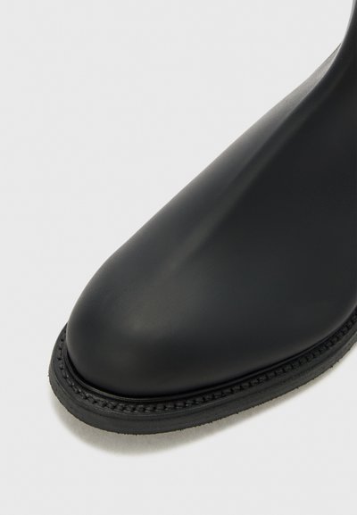 Svart ankelboot med en slät, blank yta, rund tå och en vävd kant längs kanten. Tillverkad av gummi-liknande material.