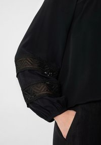 Schwarze Bluse mit voluminösen Ärmeln und komplizierten schwarzen Spitzenakzenten an den Bündchen. Schmuses Material, lockere Passform und elastische Ärmelabschlüsse.