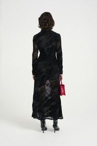 Černé dlouhé šaty s texturovanou průhlednou tkaninou, přiléhavým střihem a vysokým výstřihem, doplněné malou červenou kabelkou a černými koženými botami na podpatku.