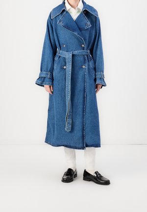 Personne portant un long trench-coat en jean bleu avec une ceinture, un pantalon blanc et des mocassins noirs, debout devant un fond blanc uni.