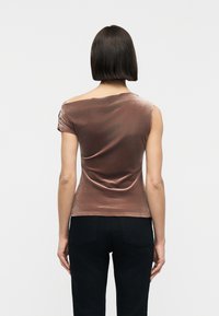 Femme aux cheveux lisses et mi-longs, vêtue d'un haut en velours marron décolleté épaules et d'un pantalon noir, debout de dos face à l'appareil photo.