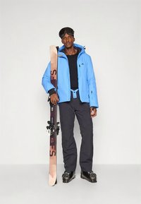 Σετ χειμερινών ρούχων για σκι που περιλαμβάνει μπλε μονωμένο μπουφάν, μαύρο εσωτερικό στρώμα, μαύρο παντελόνι για σκι και γυαλιά χιονιού. Κρατάει μπεζ σκι με μαύρες δεσίματα.