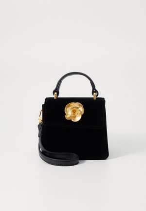 Zwarte fluwelen handtas met een gouden bloemvormige sluiting, een bovenhandgreep en een afneembare schouderriem tegen een effen witte achtergrond.