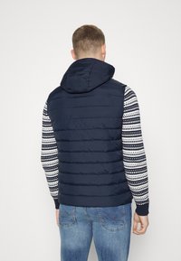 Marineblaue Steppweste mit Kapuze, mit horizontalen Nähten. Getragen über einem gemusterten Langarmshirt, kombiniert mit blauen Jeans.