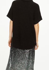 Musta neulepusero, jossa on leveä mitoitus ja lyhyet hihat, kerroksittain hopeisen paljettihameen päällä, luoden tekstuurista kontrastia.