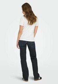 Vrouw met lang bruin haar, met de rug naar de camera gekeerd, in een eenvoudig wit T-shirt, donkerblauwe jeans en zwart-witte schoenen.