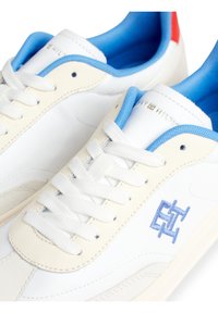 Bílé kožené tenisky s modrým límcem a tkaničkami, s modrým vyšívaným logem a červeným akcentem na patě. Hladký povrch.