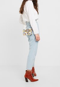 Barevná vzorovaná kabelka přes rameno s řetízkovým popruhem, nošená na boku, v kombinaci s světle modrými džíny a červenými kotníkovými botami s blokovým podpatkem.