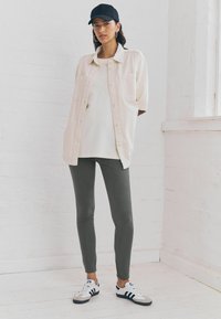 Lichtbeige overhemd over een wit T-shirt, donkergroene legging, zwarte pet en witte sneakers met blauwe strepen, staand tegen een witte muur.